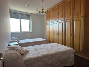 Une chambre avec deux lits, une fenêtre avec des stores, une armoire en bois et un lustre.