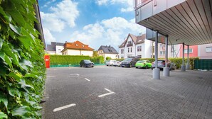 A parking area with cars, a building with a balcony, and a hedge. - Hotel Reichshof (Schwerte)