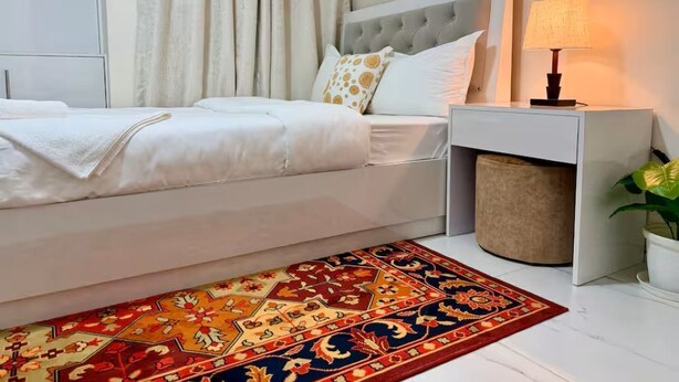 A bedroom with a bed, a bedside table with a lamp, a rug, and a plant.
