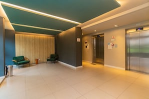 A hotel lobby with a modern design, featuring a reception desk, two green chairs, an elevator, and a trash bin. - Apto com Smart Lock Perto do Aeroporto e Metr (São Paulo)