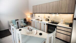 A modern kitchen with a built-in oven, a coffee maker, and a sink. There is a dining area with a glass table and white chairs.