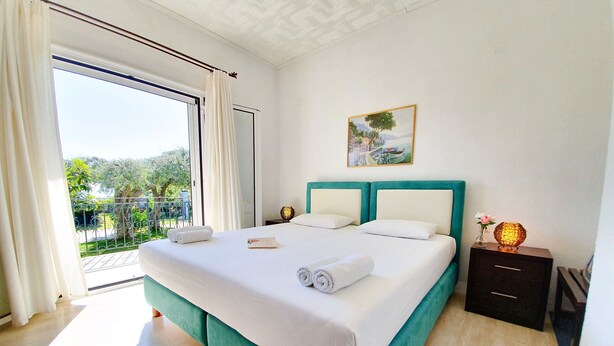 A hotel room with a large bed, a view of the outdoors, a painting on the wall, and a nightstand with a lamp. - Double room on ground floor (Syvota)