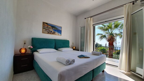 Un dormitorio con cama, mesita de noche, un cuadro y balcón con una palmera.