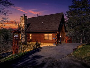 Une cabane en rondins avec une cheminée en pierre, une véranda et une allée menant à l’entrée.