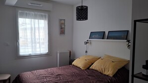 Une chambre à coucher avec un lit, une fenêtre avec des rideaux, un radiateur, une étagère avec des photos et un luminaire suspendu.