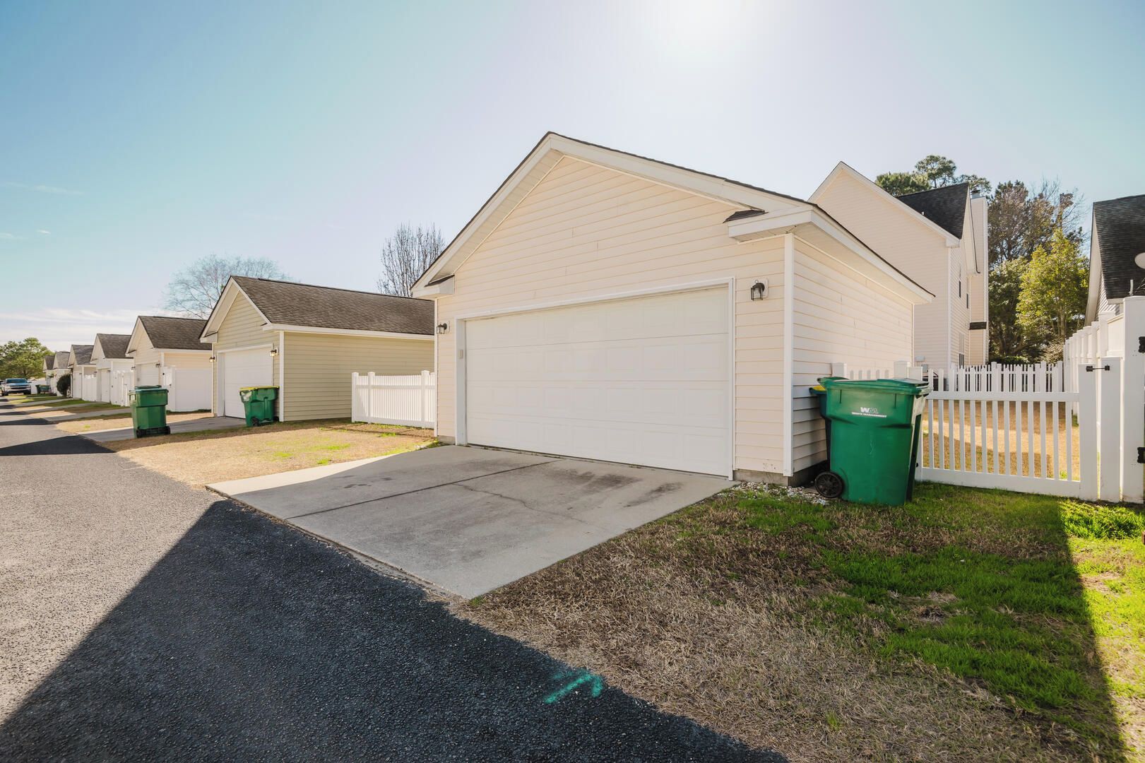 A row of single-story houses with attached garages, white picket fences, and green trash bins.