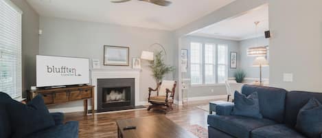 A living room with a fireplace, a television, a ceiling fan, and a large window.