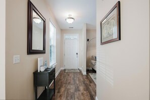 A hallway with wooden flooring, a white door, a black console table with items, and a framed picture on the wall. - Dogs Welcome! Adorable 3-bedroom Coastal Home in Historic Old Town Bluffton (Bluffton)