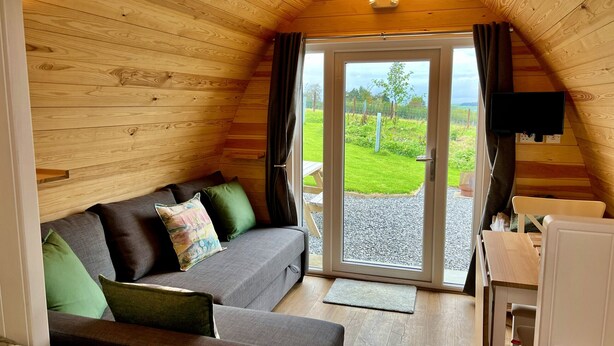 En mysig stuga med träpanel i taket, en soffa med kuddar, en tv som är monterad på väggen och en skjutdörr i glas som leder ut till ett område utomhus.