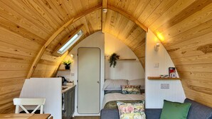 Ett mysigt, kompakt rum med ett sluttande tak, en liten köksdel, en säng med randigt sänglinne och en soffa med en grön kudde.