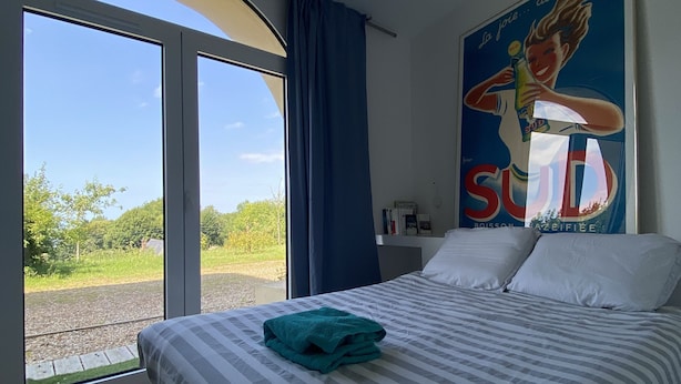 A bedroom with a large bed, a window with curtains, a poster on the wall, and a view of greenery outside. - WHOLE APARTMENT WITH PARK AND SEA VIEW TROUVILLE SUR MER. (Trouville-sur-Mer)