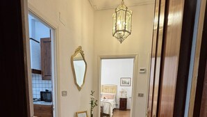 Un couloir doté d’un plancher en bois, d’un miroir, d’un lustre et d’une porte donnant sur une chambre à coucher avec un lit et une table de chevet.