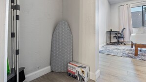 Un angolo di stanza con pavimento in legno, parete bianca, una tavola da stiro grigia, una sedia da ufficio nera, un tavolino con una lampada e un letto con copripiumino bianco.