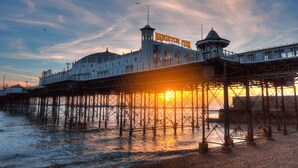 A pier with a lighthouse and a building labeled 'Brighton Pier' under a sunset sky. - Oriental Three | By My Getaways (Brighton)
