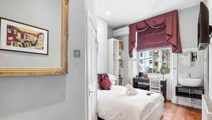A bedroom with a bed, a window with curtains, a bathroom with a sink, and a framed picture on the wall. - Oriental Three | By My Getaways (Brighton)