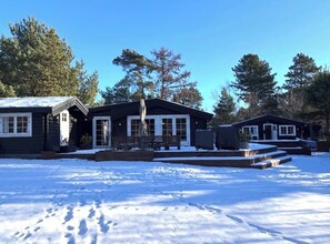 A cabin with a snow-covered ground, a wooden deck, and a large window.