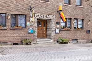 A brick building with a sign reading 'GASTHAUS' above the entrance, potted plants on the steps, and a flag.
