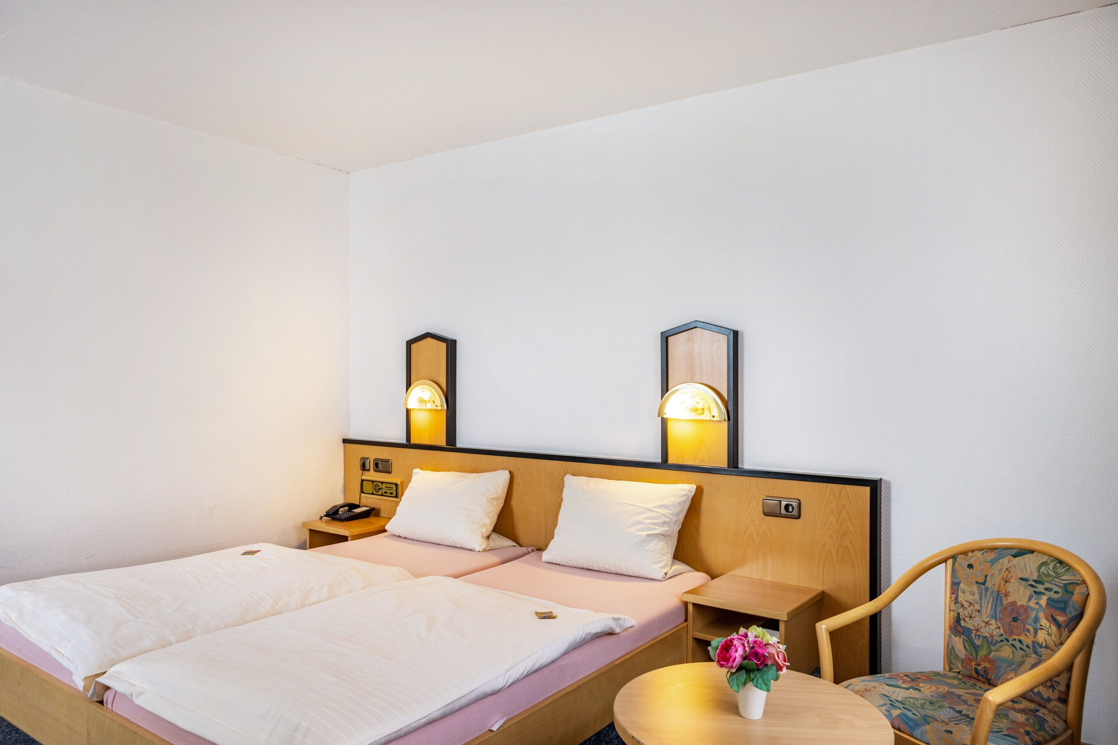 A hotel room with two single beds, bedside tables with lamps, a wooden headboard, a round wooden table, and a floral chair.