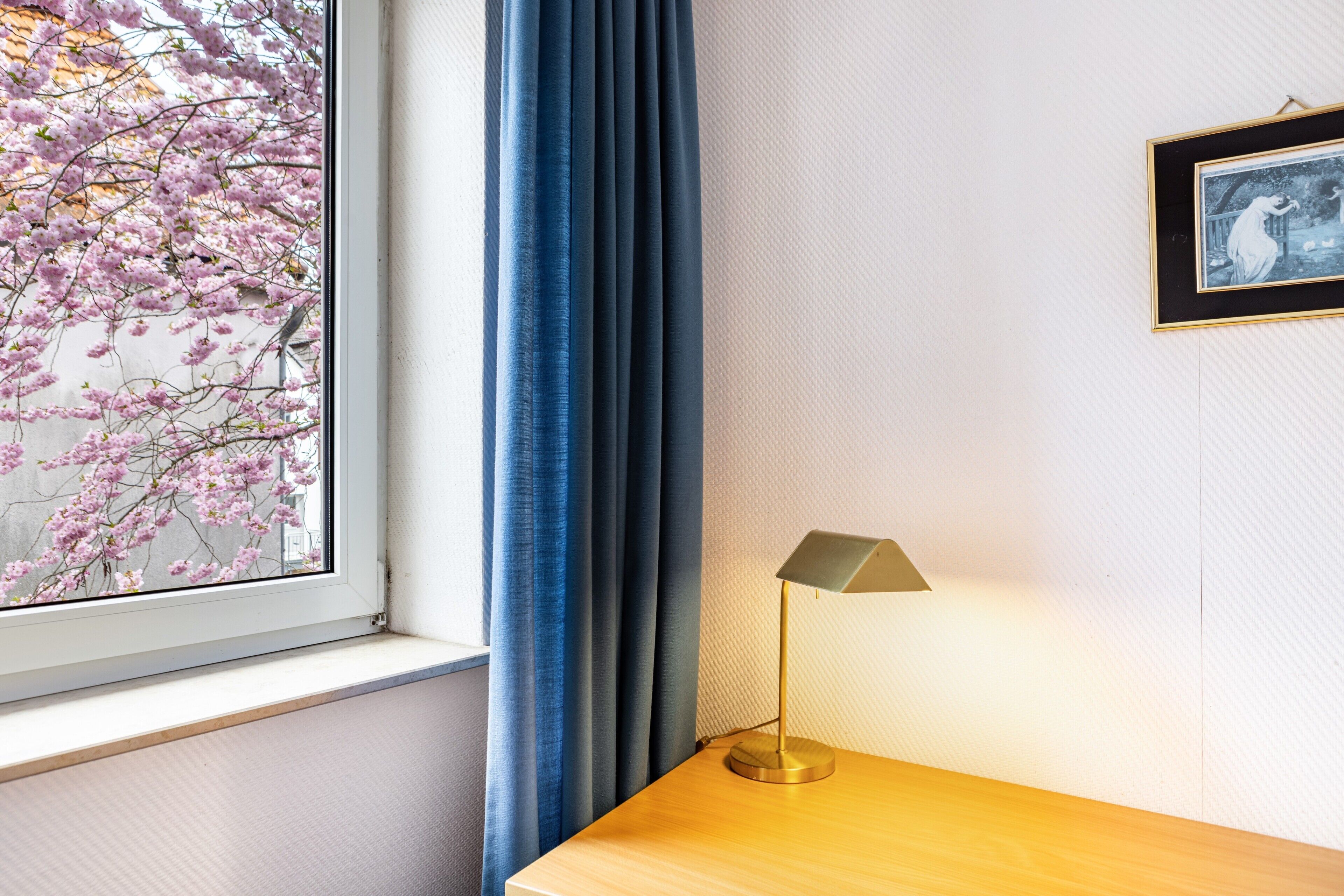 Une pièce avec une fenêtre, une lampe posée sur une table en bois et un tableau encadré accroché au mur.