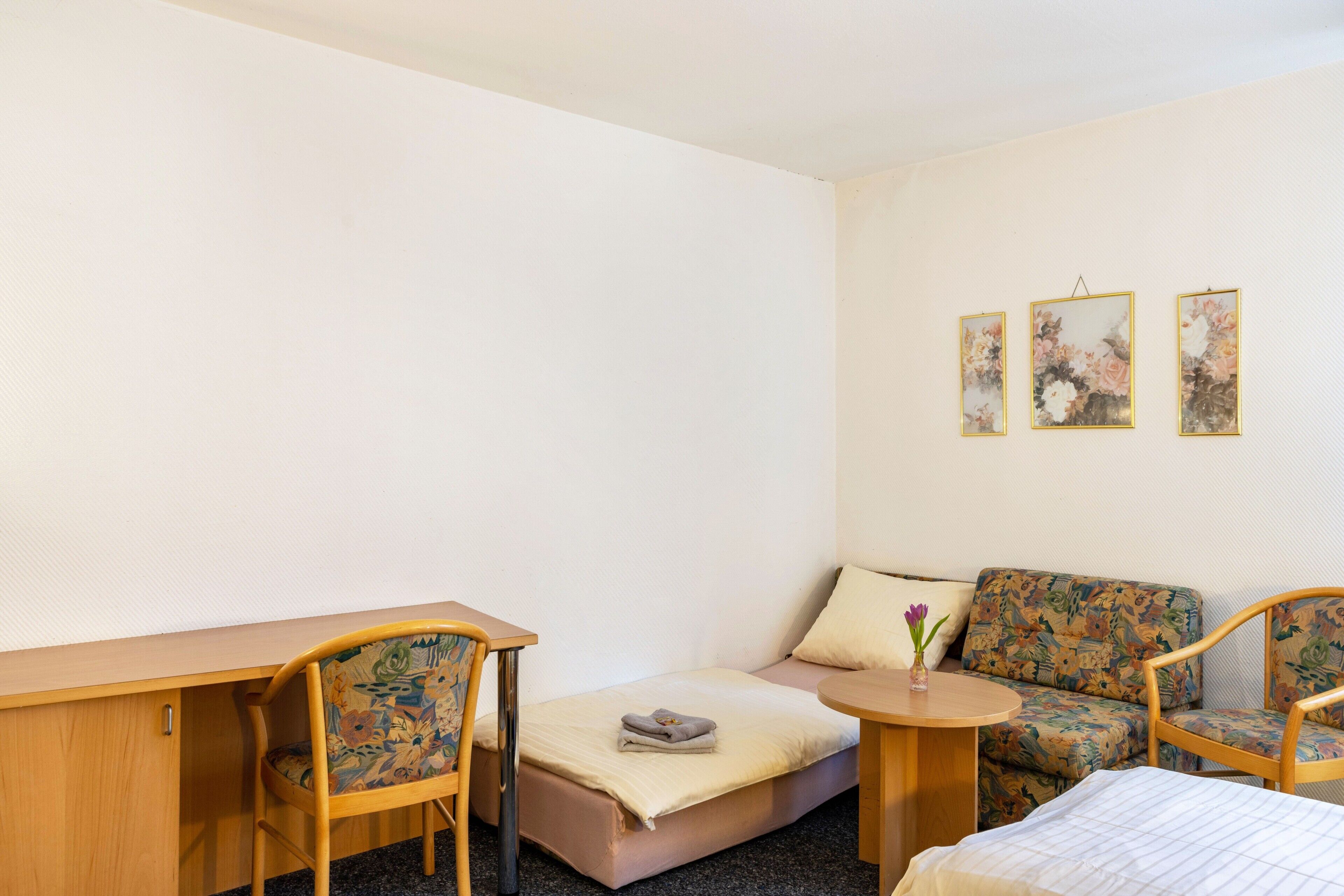 Una habitación pequeña con una cama, un escritorio, una silla, un sofá y tres cuadros colgados en la pared.