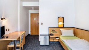 Habitación de hotel con escritorio, silla, cama, mesita de noche, lámpara y un pequeño refrigerador.