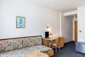 Ein Zimmer mit einer Couch, einem Schreibtisch, einem Sessel, einem kleinen Tisch mit einer Vase und einem Kühlschrank.
