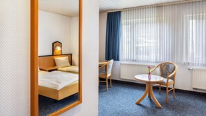 Ett hotellrum med en säng, en stol, ett litet bord, en spegel och ett fönster med gardiner.