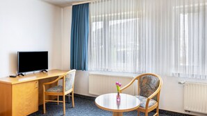 Ett hotellrum med ett skrivbord i trä, två stolar, ett litet bord med en vas, en tv och stora fönster med tunna, genomskinliga gardiner.