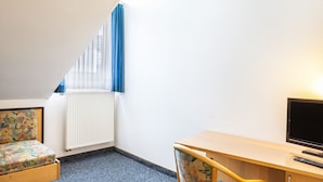 Ein kleiner, gut beleuchteter Raum mit einem Bett, einem Schreibtisch, einem Stuhl und einem Fernseher.