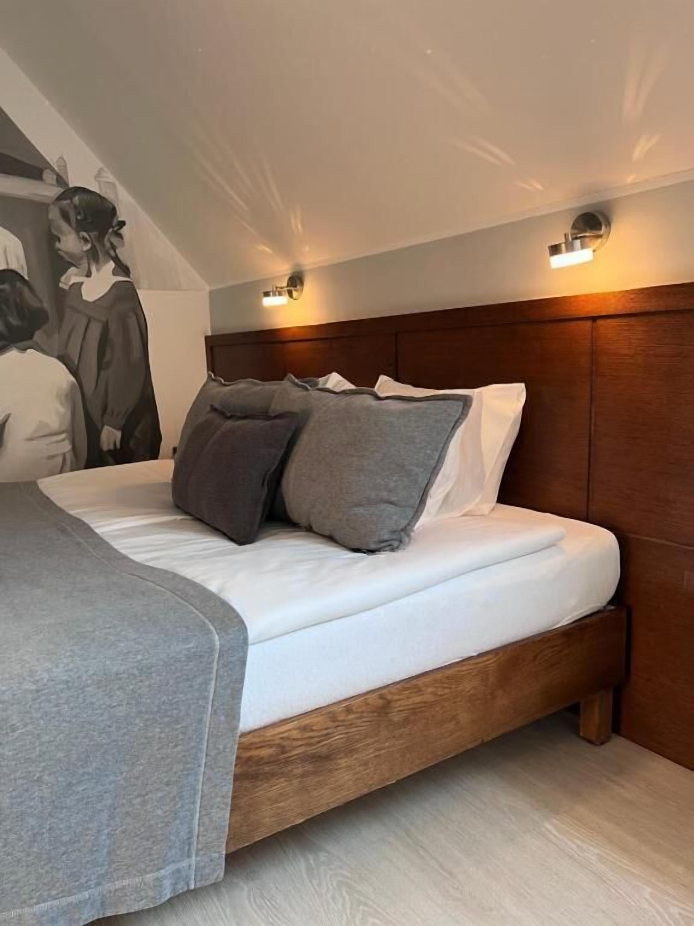 A modern bedroom with a wooden headboard, a large bed with grey and white bedding, and a wall-mounted light fixture.