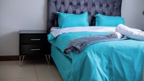 Ein ordentlich bezogenes Bett mit grauem Kopfteil, einem schwarzen Nachttisch mit Schubladen und einer türkis-farbenen Bettdecke.
