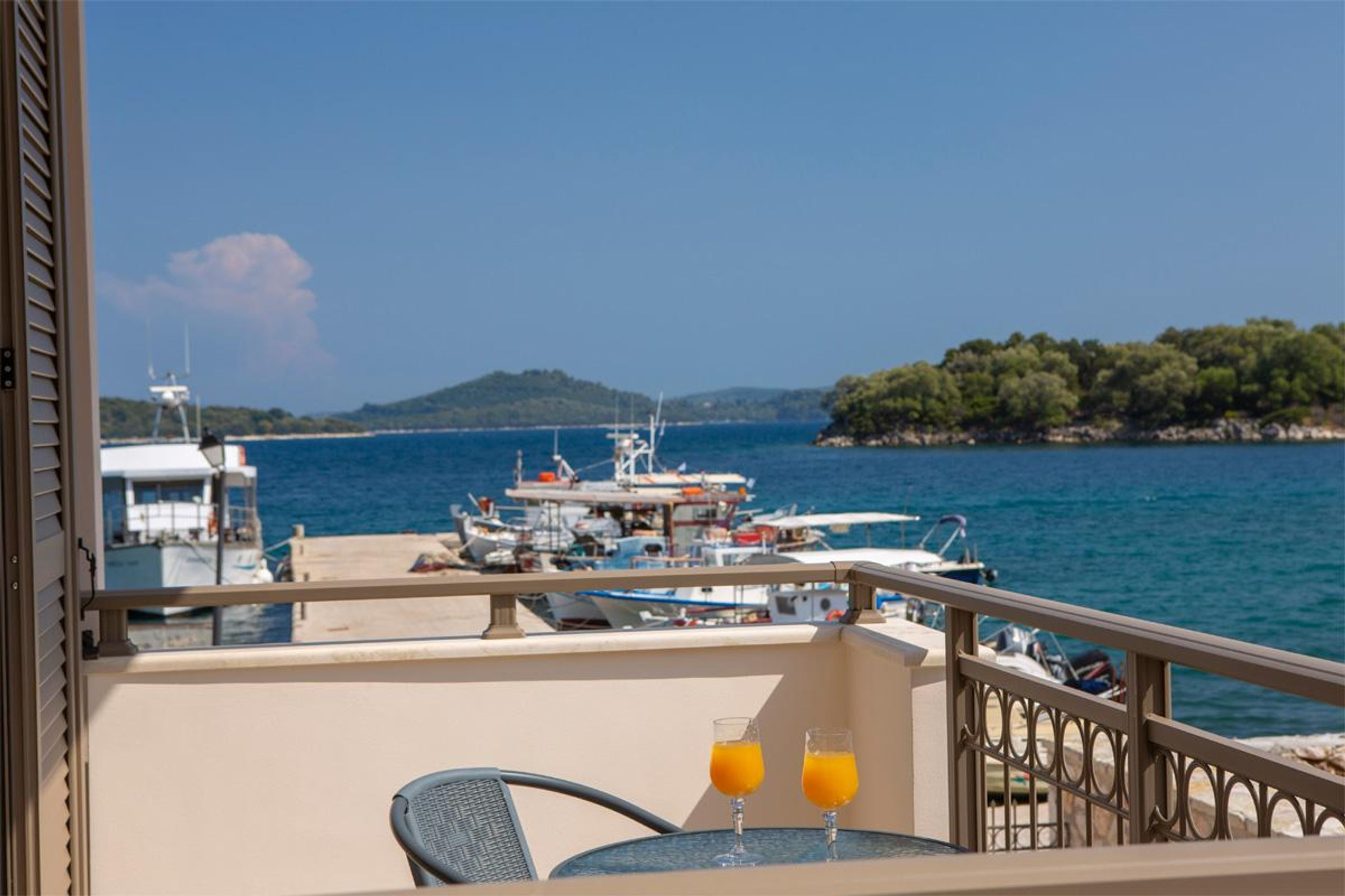 A balcony with a table and two glasses of orange juice, overlooking a marina with boats and a hilly landscape.