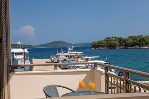Un balcon avec une table et deux verres de jus d’orange, donnant sur une marina où sont amarrés des bateaux, et sur un paysage vallonné.