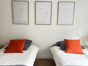 Two single beds with white bedding and orange pillows, a white wall, and three framed artworks above the beds. - 2 bedroom apartment - Downtown Parthenay (Parthenay)
