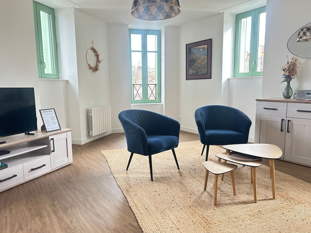 A modern living room with a TV, two blue armchairs, a small wooden table, and a view through a window.