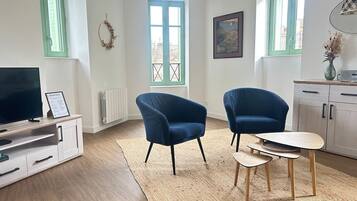 A modern living room with a TV, two blue armchairs, a small wooden table, and a view through a window.