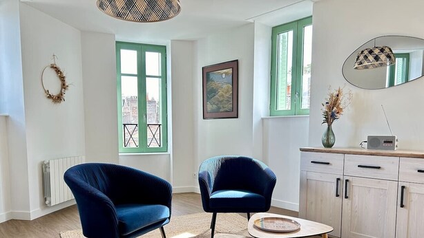 Una stanza con due poltrone blu, un tavolino rotondo, un mobile con cassetti, un termosifone e una finestra con cornice verde.