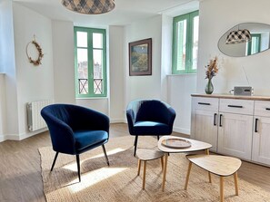A room with two blue armchairs, a small round table, a cabinet with drawers, a radiator, and a window with a green frame. - 2 bedroom apartment - Downtown Parthenay (Parthenay)
