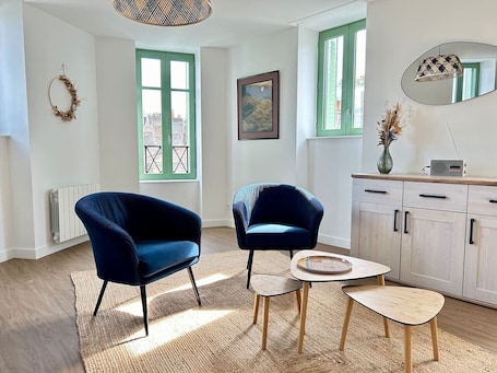 A room with two blue armchairs, a small round table, a cabinet with drawers, a radiator, and a window with a green frame.. 2 bedroom apartment - Downtown Parthenay