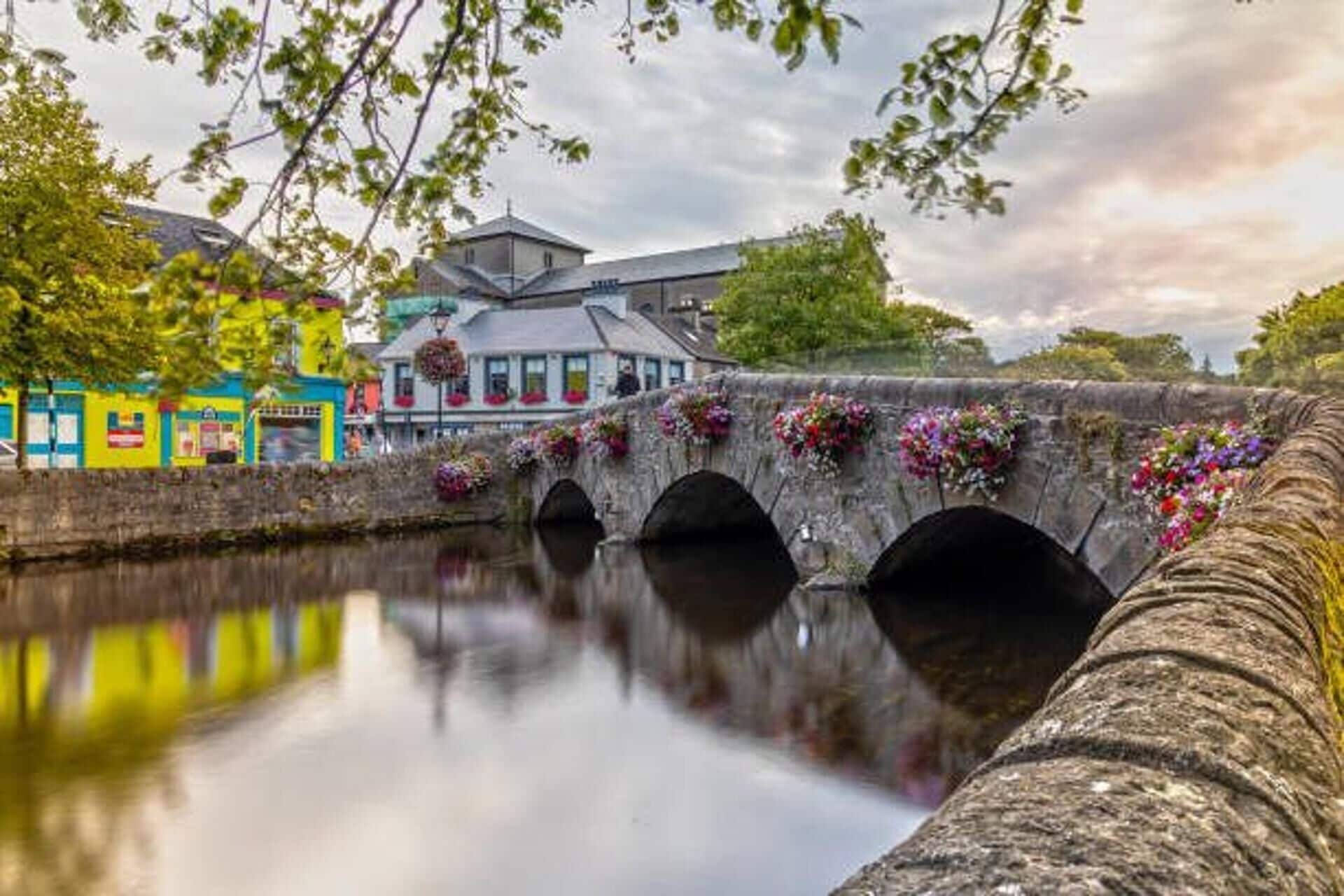 A stone bridge with flower baskets, a river, and buildings with colorful facades.