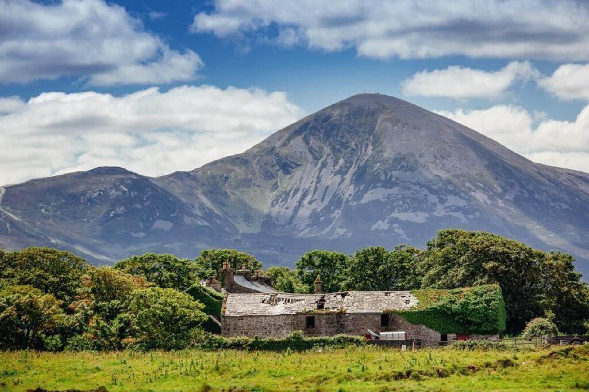 A stone building with a sloped roof, surrounded by greenery and a mountain in the background.