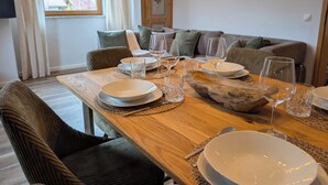 A dining area with a wooden table set for four, surrounded by upholstered chairs, a sofa, and a window with curtains. - Chic Alpine Oasis Oberammergau with Mountain View (Oberammergau)
