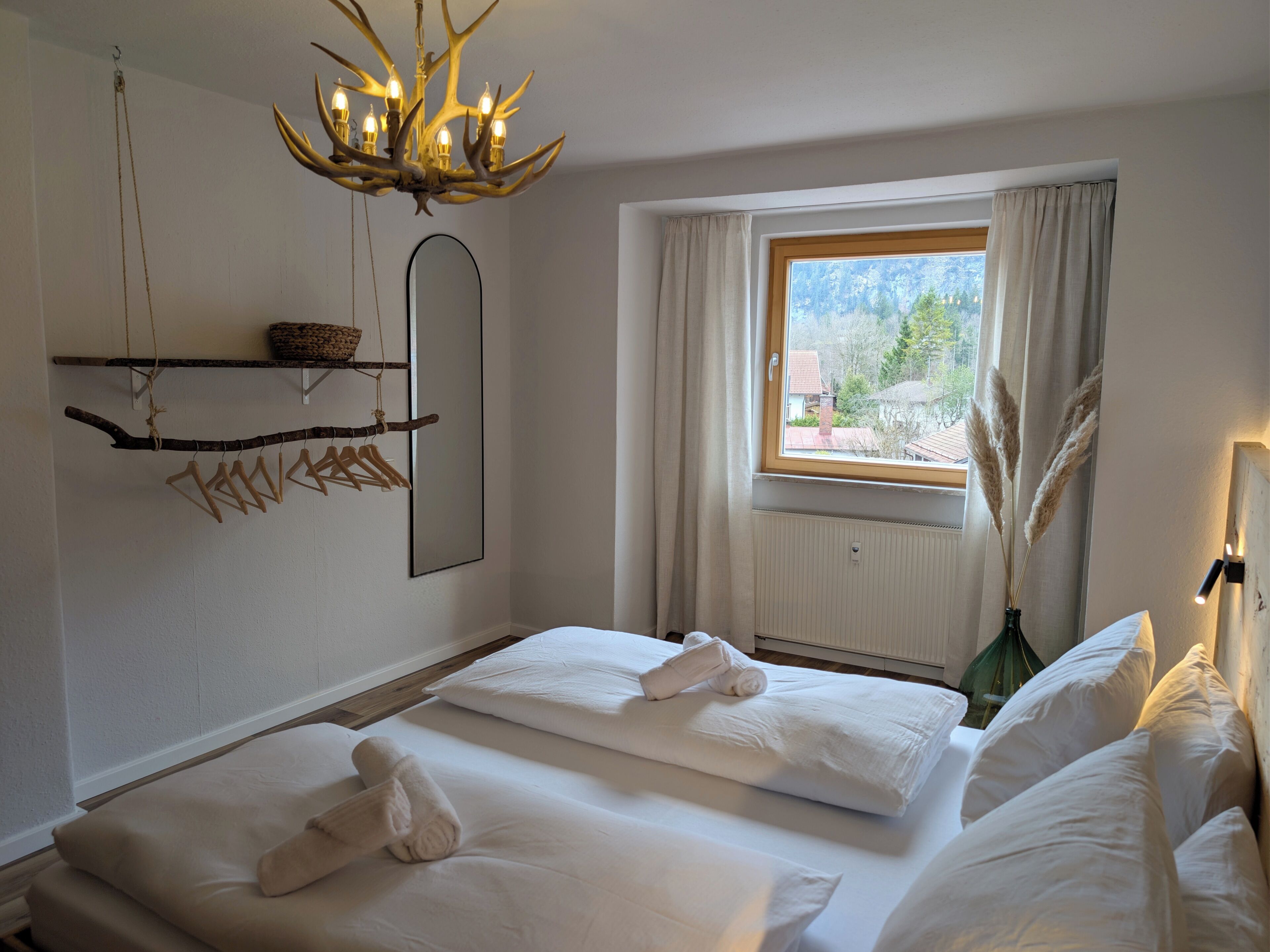 A bedroom with two beds, a chandelier, a window with curtains, a mirror, and a hanging branch with clothes.