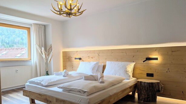 A bedroom with a wooden bed, a chandelier, a window with curtains, and a wooden headboard. - Chic Alpine Oasis Oberammergau with Mountain View (Oberammergau)