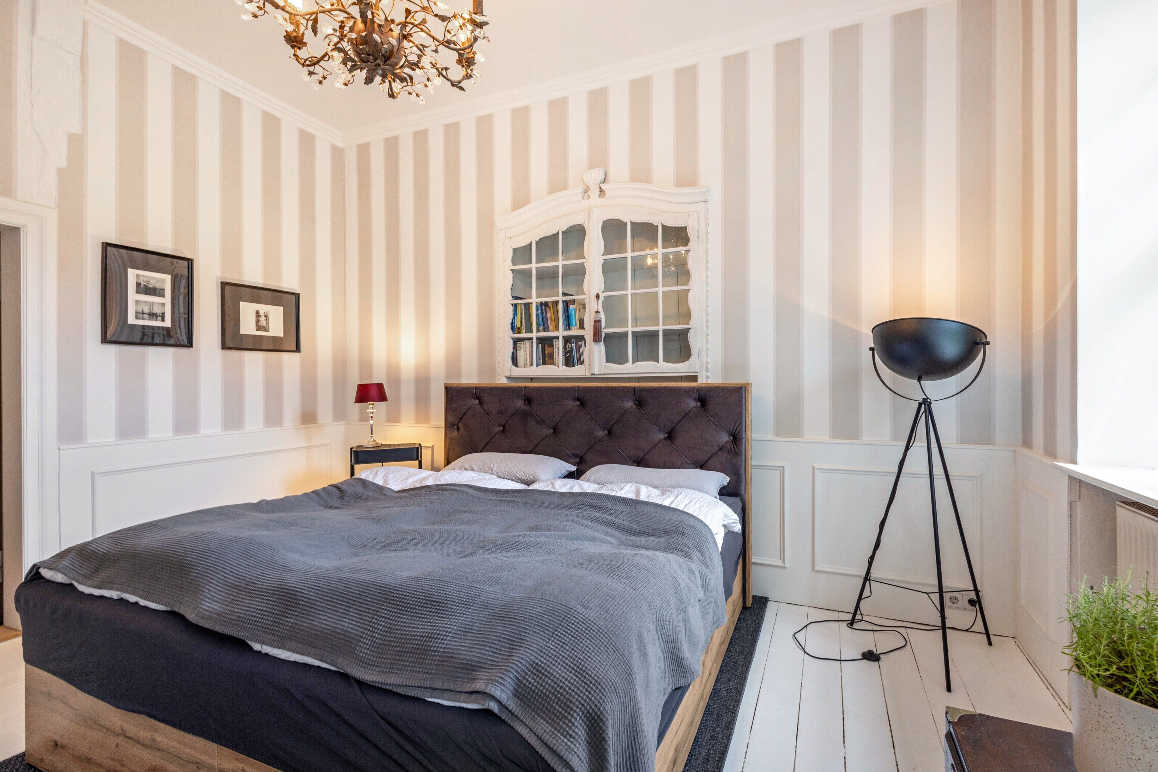 Ein Schlafzimmer mit einem großen Bett, einem Nachttisch, einer Stehlampe, einem Fenster mit Bücherregalen und einer Kristallleuchter.