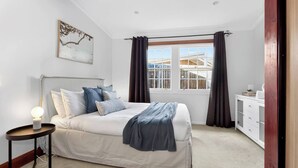 Een slaapkamer met een bed, een nachtkastje met een lamp, een raam met gordijnen en een witte dressoir.