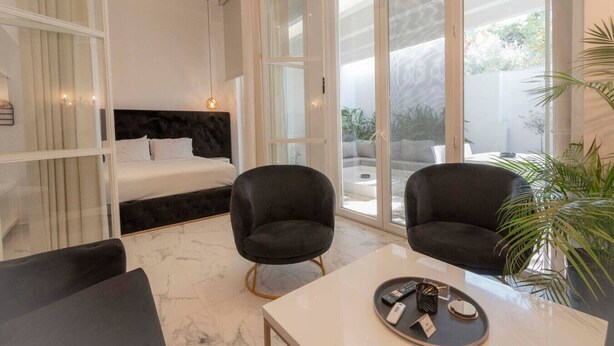 A modern hotel room with a large bed, two armchairs, a small table, and a view of the outdoors. - 4 Elemental Living Suites - Wind (Nea Moudania)