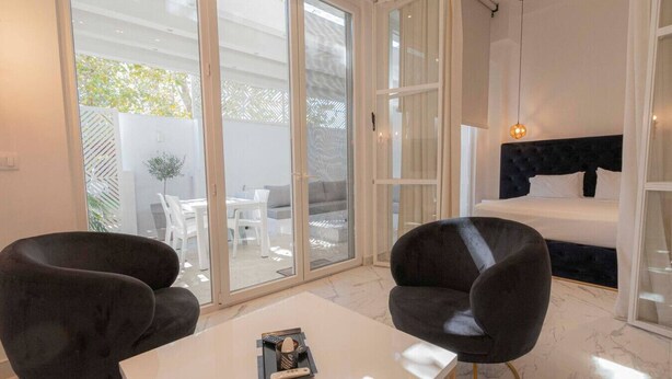Ein modernes Schlafzimmer mit einem Bett, zwei Sesseln, einem kleinen Tisch und einer Glasschiebetür, die zu einem Außenbereich führt.