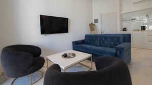 Un salon comprenant un canapé, deux fauteuils, une table basse, un téléviseur à écran plat et un coin cuisine avec des placards et une machine à café.