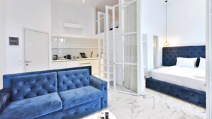 Une chambre d’hôtel moderne avec un canapé bleu, un lit doté d’une tête de lit sombre, une grande fenêtre et un coin cuisine à l’arrière-plan.